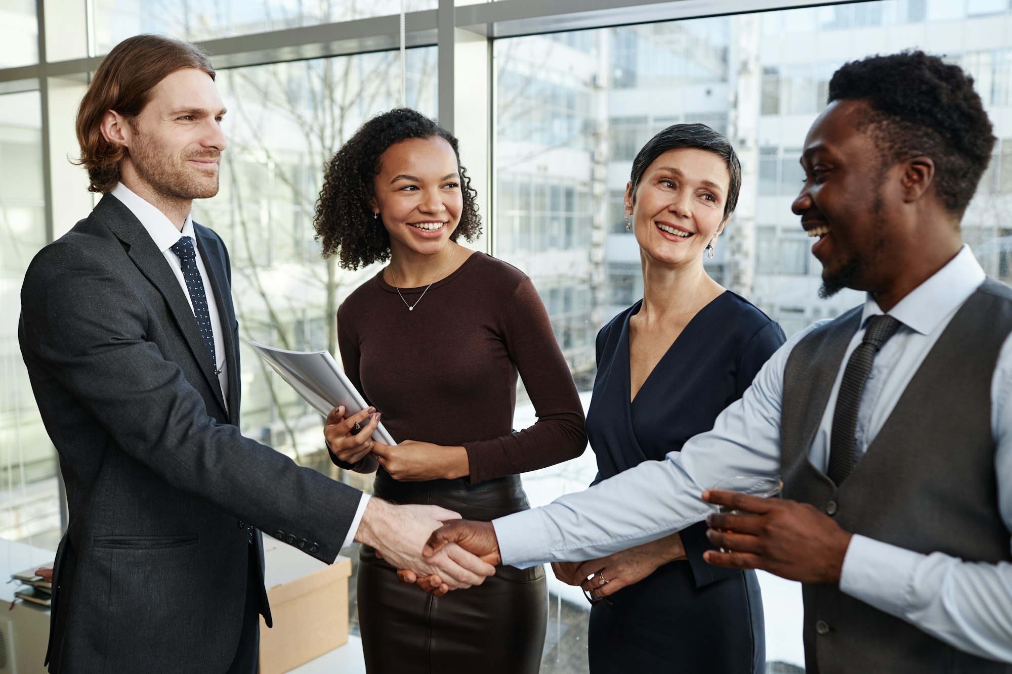 Smiling business partners shaking hands in office standing with drinks